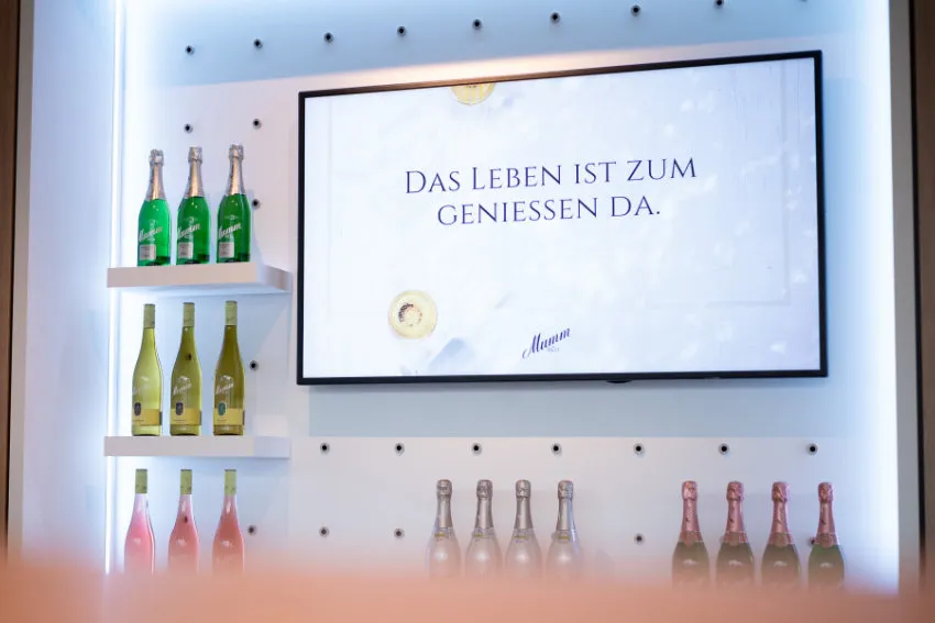 Bildschirm mit Markenbotschaft zwischen Sektflaschen von Rotkäppchen-Mumm